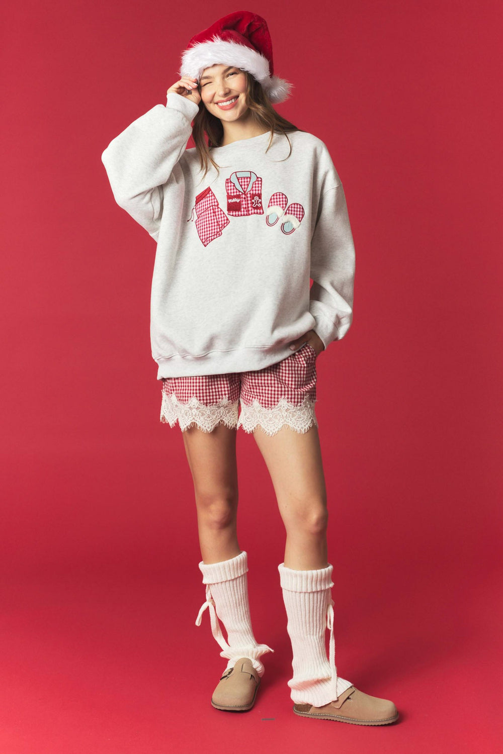 A woman modeling a cozy Christmas outfit consisting of a light gray sweatshirt with a red and white holiday graphic, red and white gingham shorts with a white lace hem, a red Santa hat, and beige slip-on shoes, set against a solid red studio background.