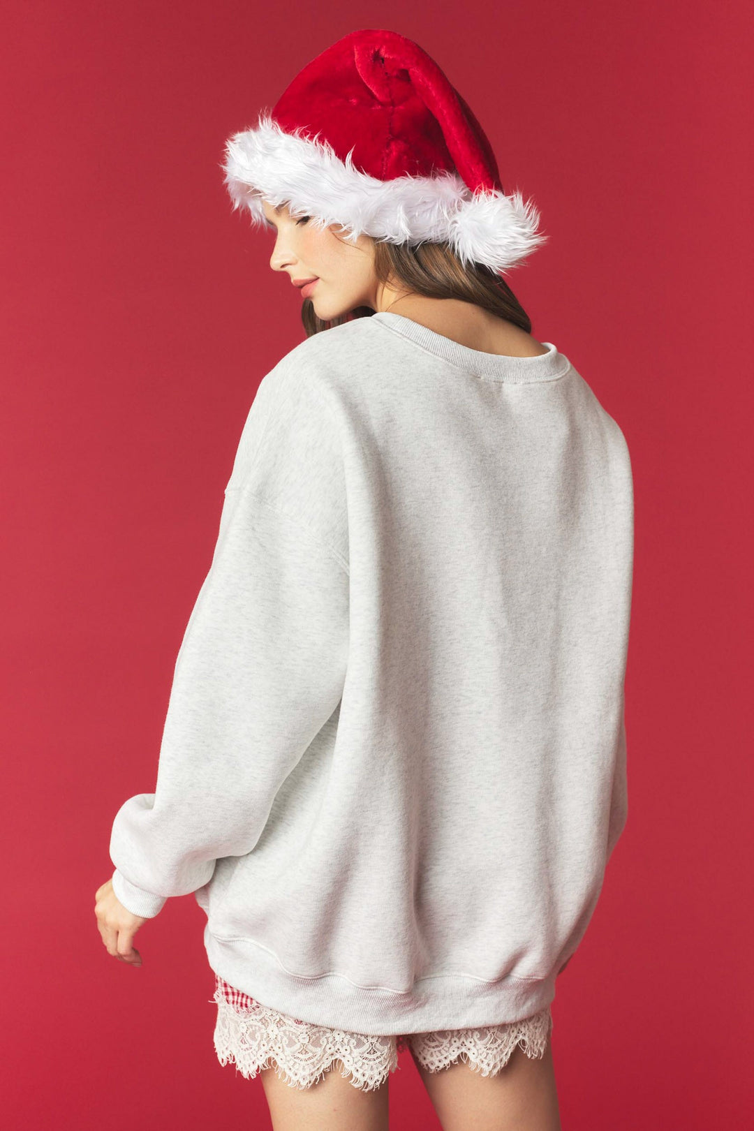 A woman wearing an oversized light gray crewneck sweatshirt, a red Santa hat with white trim, and lace-trimmed shorts, photographed against a solid red studio background.