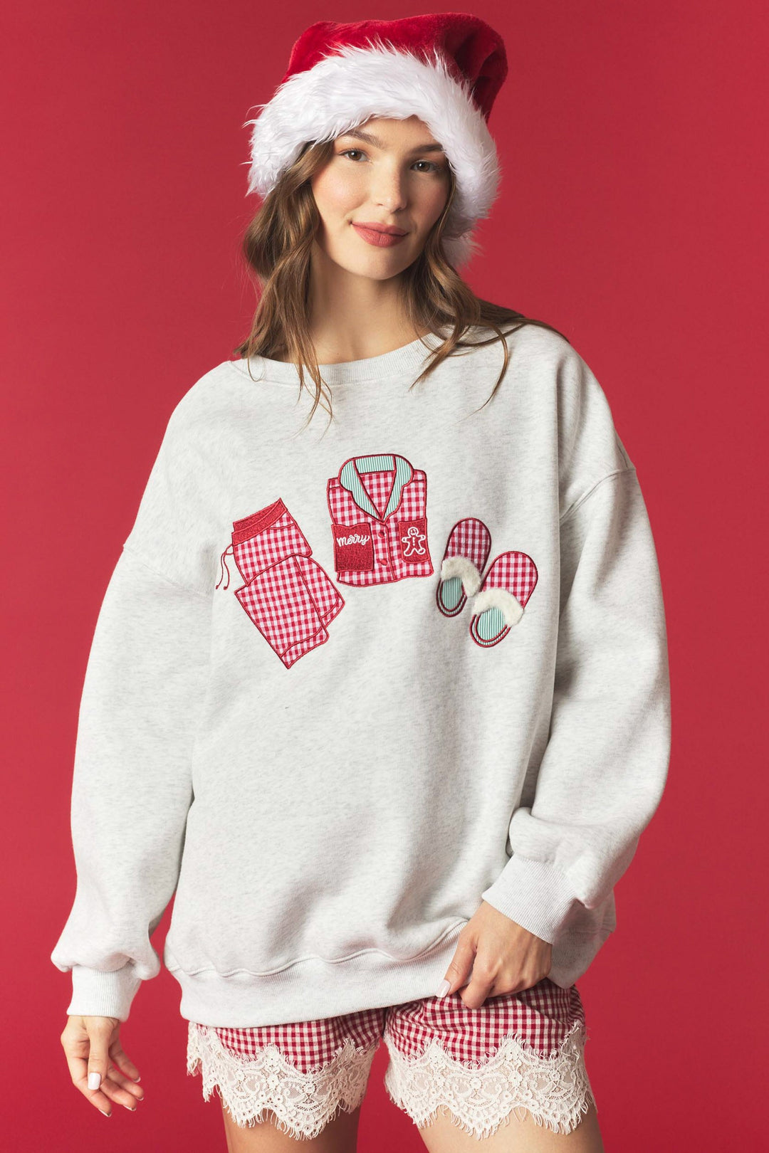 A woman wearing a light gray crewneck sweatshirt with a red and white gingham pajama-themed graphic on the front, styled with matching red and white gingham pajama shorts and a Santa hat, against a solid red background.