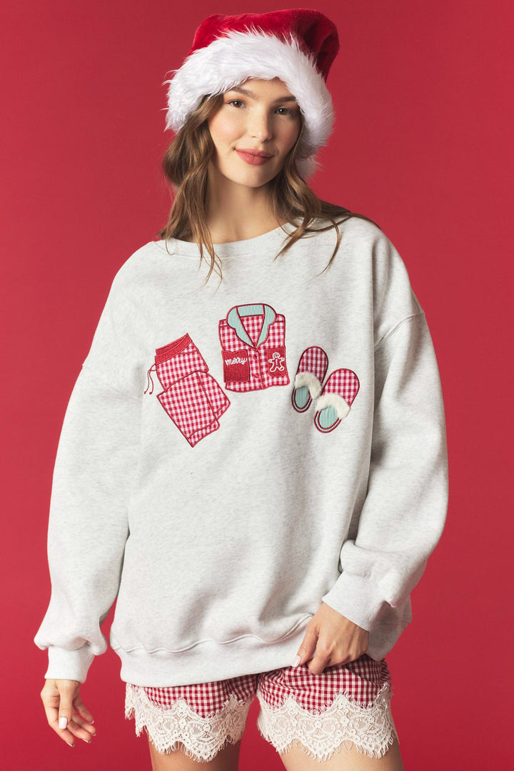 A woman wearing a light gray crewneck sweatshirt with a red and white gingham pajama-themed graphic on the front, styled with matching red and white gingham pajama shorts and a Santa hat, against a solid red background.
