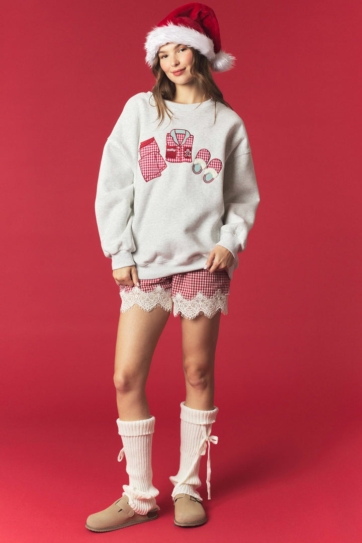 A woman modeling a cozy holiday-themed outfit consisting of a light gray sweatshirt with a red and white gingham robe graphic, matching red and white gingham shorts with white lace trim, white leg warmers, and beige slip-on shoes, complete with a red Santa hat, set against a solid red studio background.