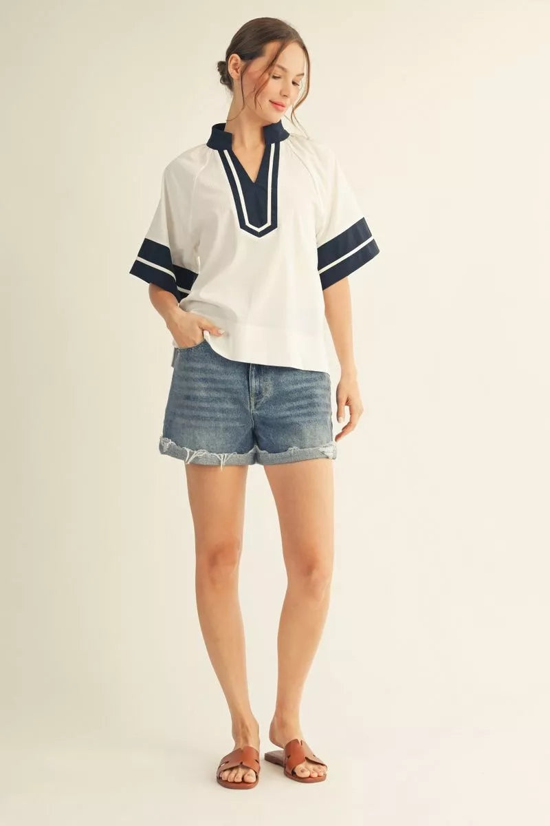 Woman wearing a white blouse with navy accents and denim shorts on a plain background