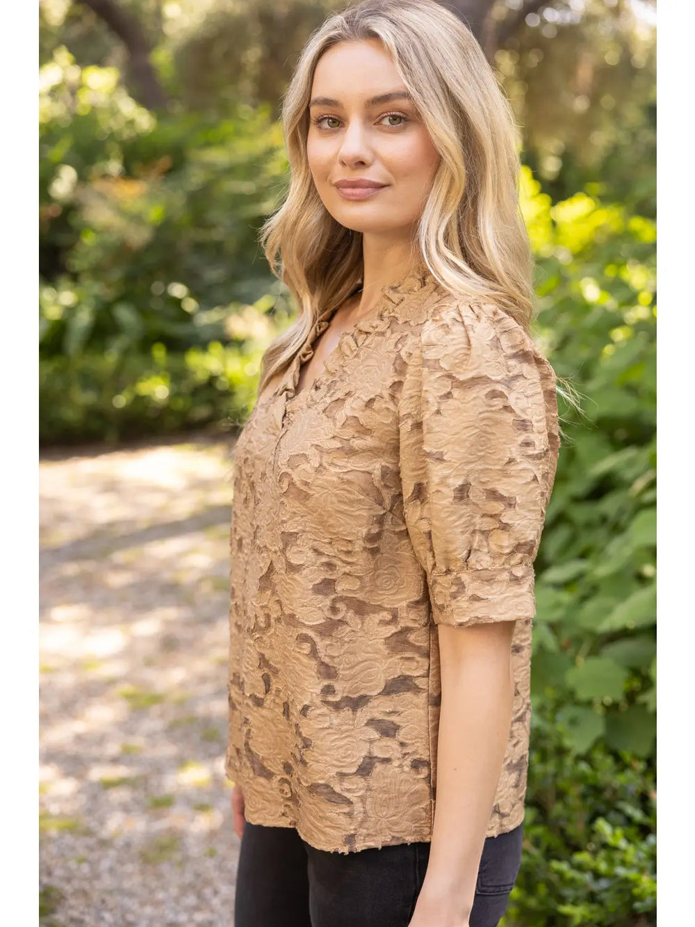 A woman wearing a tan textured lace short-sleeve top with a split neckline, standing outdoors in a garden setting.
