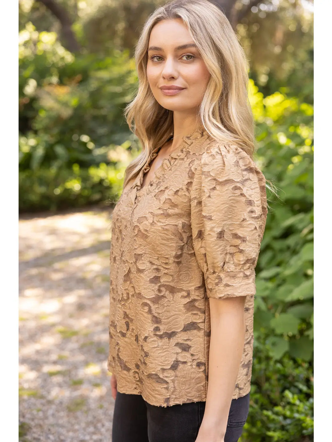 A woman wearing a tan textured lace short-sleeve top with a split neckline, standing outdoors in a garden setting.