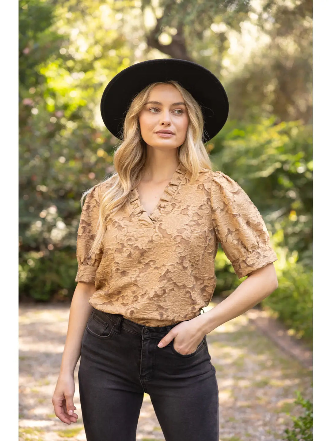 A woman wearing a tan textured short-sleeve blouse with ruffles, black jeans, and a black wide-brim hat, standing outdoors on a sunlit path with blurred green foliage in the background.