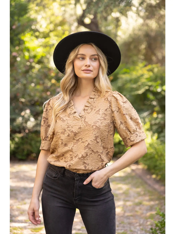 A woman wearing a tan textured short-sleeve blouse with ruffles, black jeans, and a black wide-brim hat, standing outdoors on a sunlit path with blurred green foliage in the background.