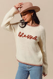 Woman wearing a cream sweater with 'blessed' text, blue jeans, and a beige hat on a beige background