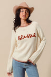 Woman wearing a cream sweater with 'blessed' text, blue jeans, and a pink hat on a beige background