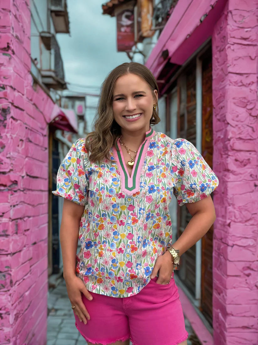 Woman in a floral blouse and pink shorts standing in a narrow alley with pink walls.
