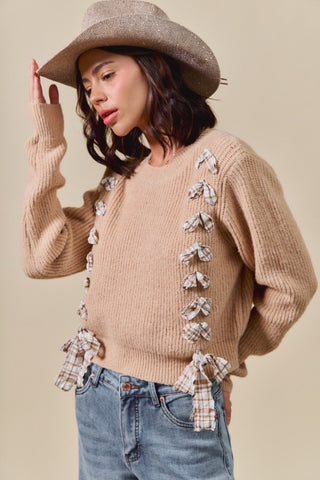 Woman wearing a beige sweater with bow details, blue jeans, and a wide-brimmed hat on a beige background