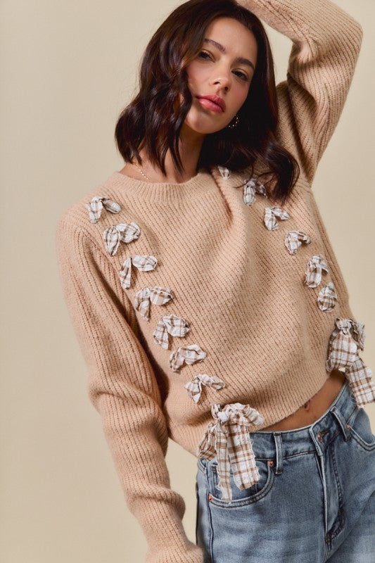 Woman wearing a beige sweater with decorative bows and plaid accents, standing against a plain background.