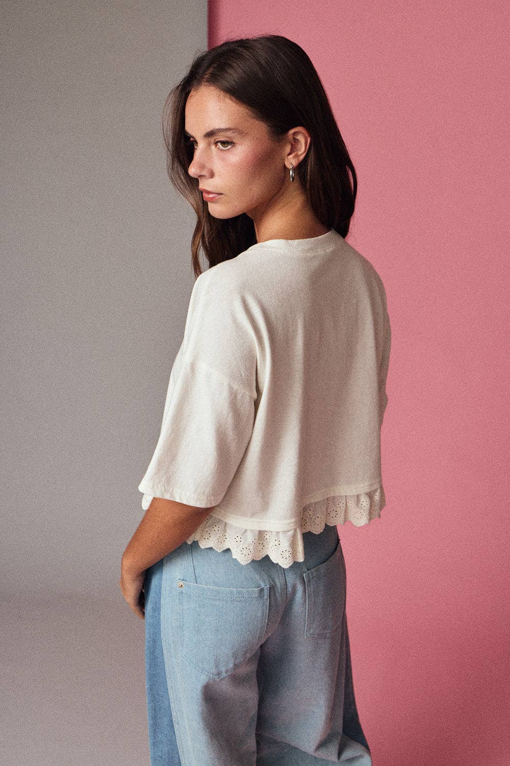 Woman wearing a white blouse with ruffled details and light blue jeans against a pink wall.