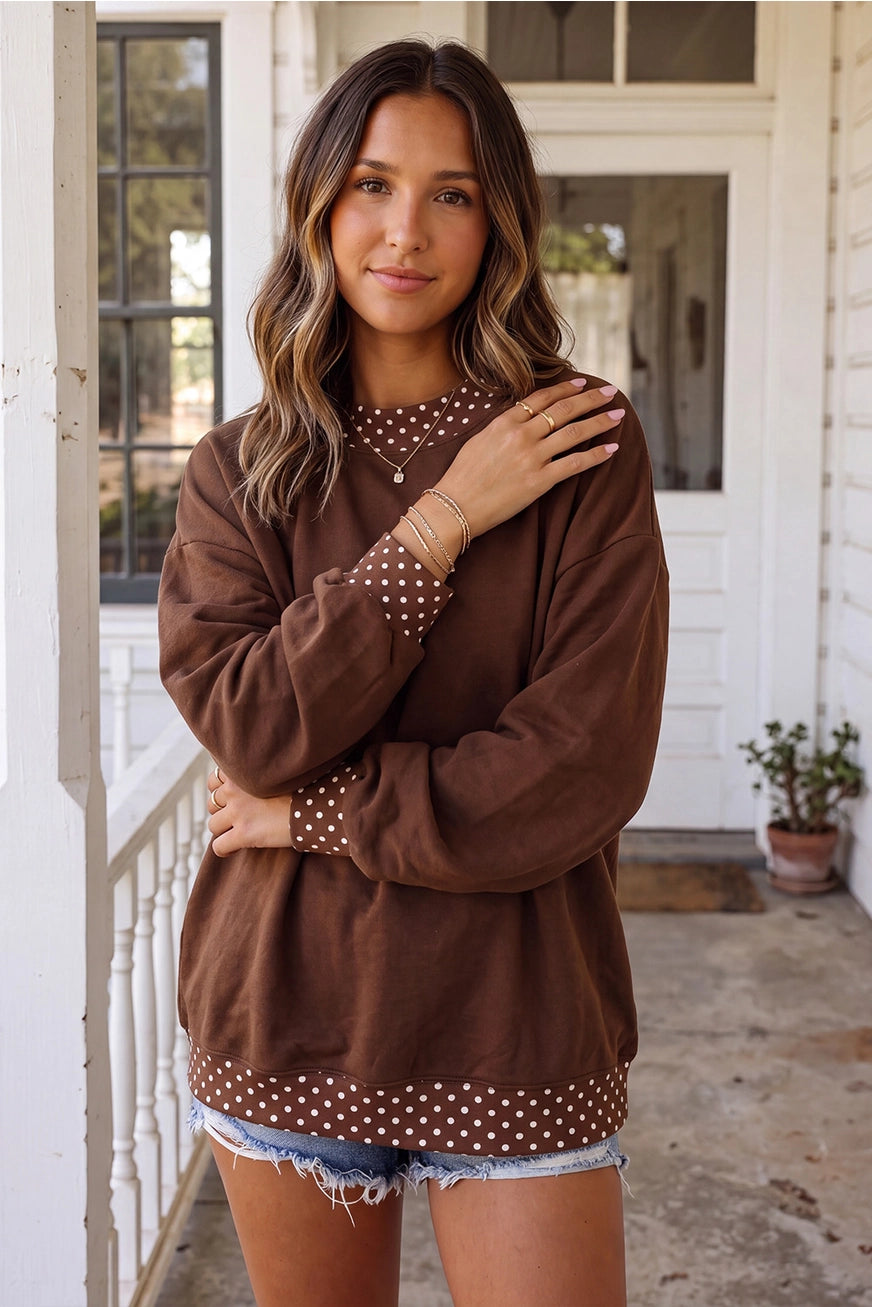 A woman modeling a brown long-sleeve sweatshirt with white polka dot trim on the collar, cuffs, and hem, paired with denim shorts, standing on a porch.
