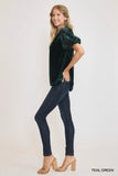 Woman wearing a teal green velvet top and dark jeans against a white background