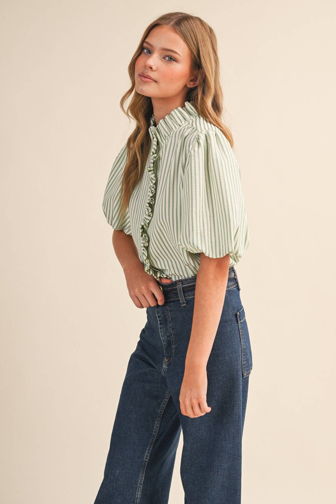 Woman wearing a green and white striped blouse with ruffled details and blue jeans on a beige background
