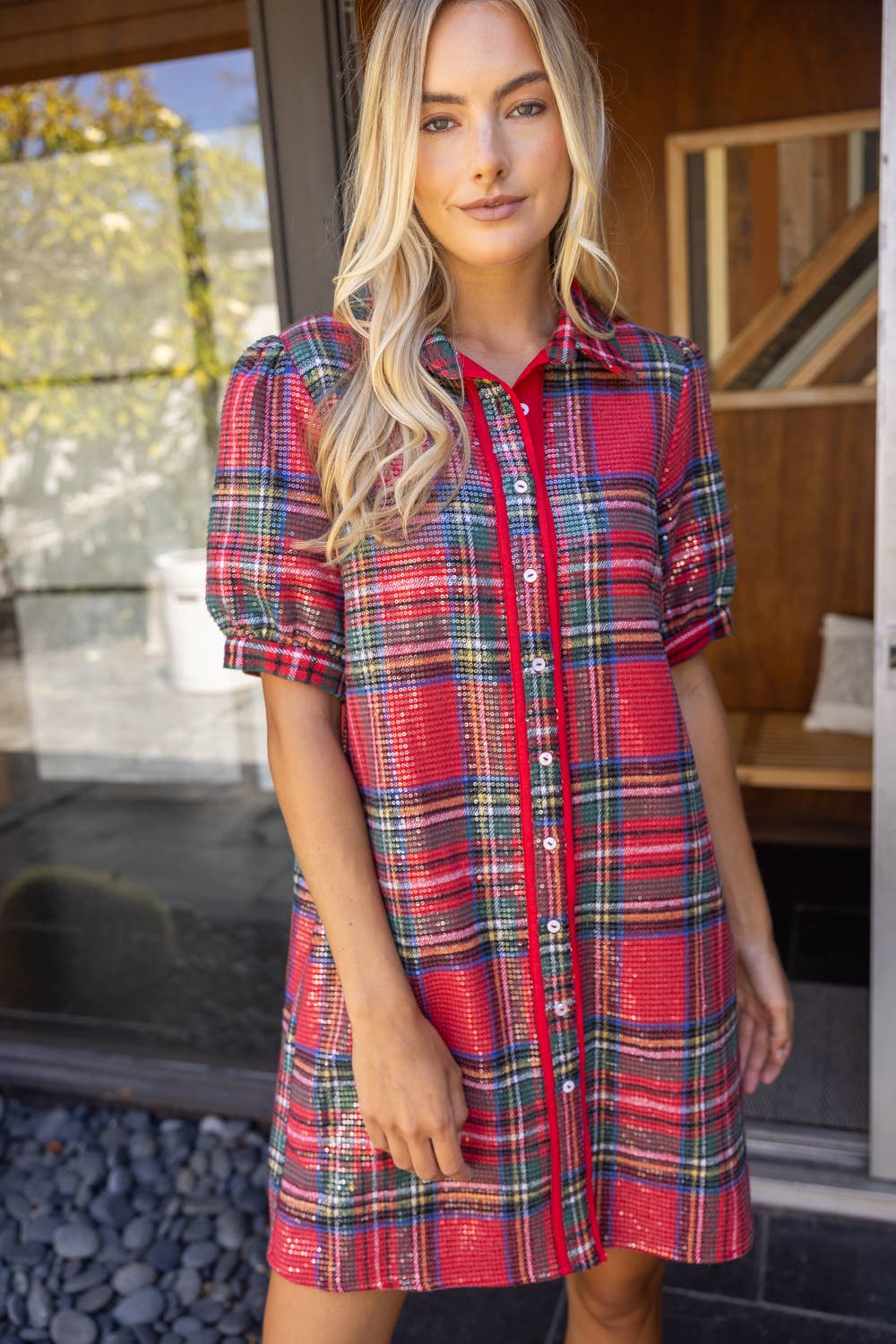 A woman wearing a red plaid short-sleeve button-up shirt dress, standing in front of a glass door and wooden wall.