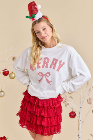 Woman wearing a 'Merry' sweater with a red bow and a Santa hat on a beige background.