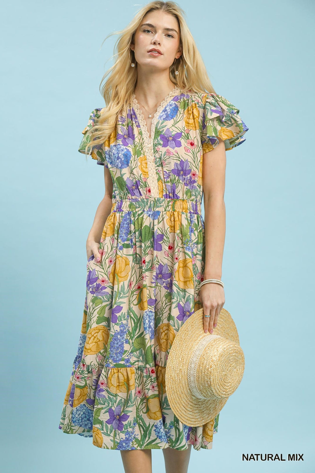 Woman wearing a colorful floral dress holding a straw hat against a light blue background