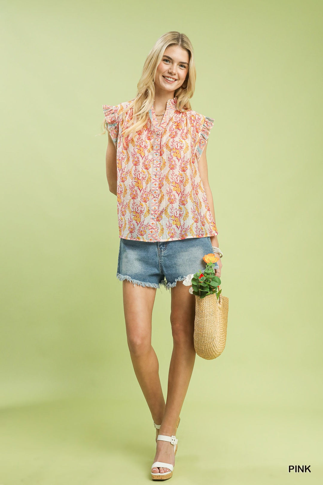Woman wearing a floral sleeveless top and denim shorts, holding a woven bag with a plant, on a light green background.