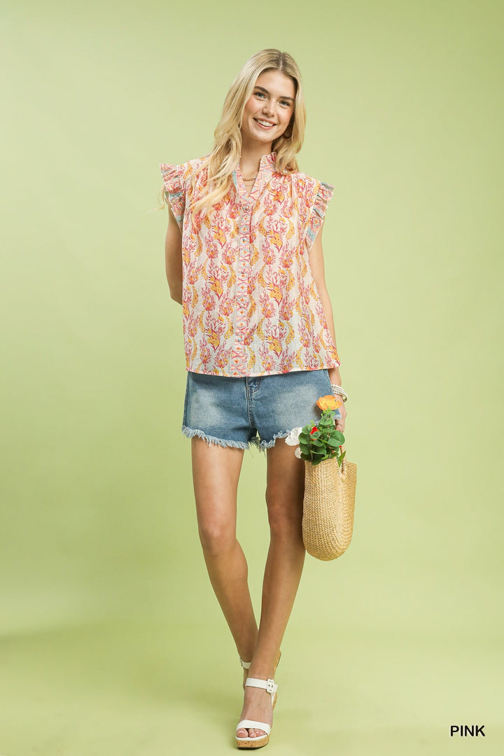 Woman wearing a floral sleeveless top and denim shorts, holding a woven bag with a plant, on a light green background.