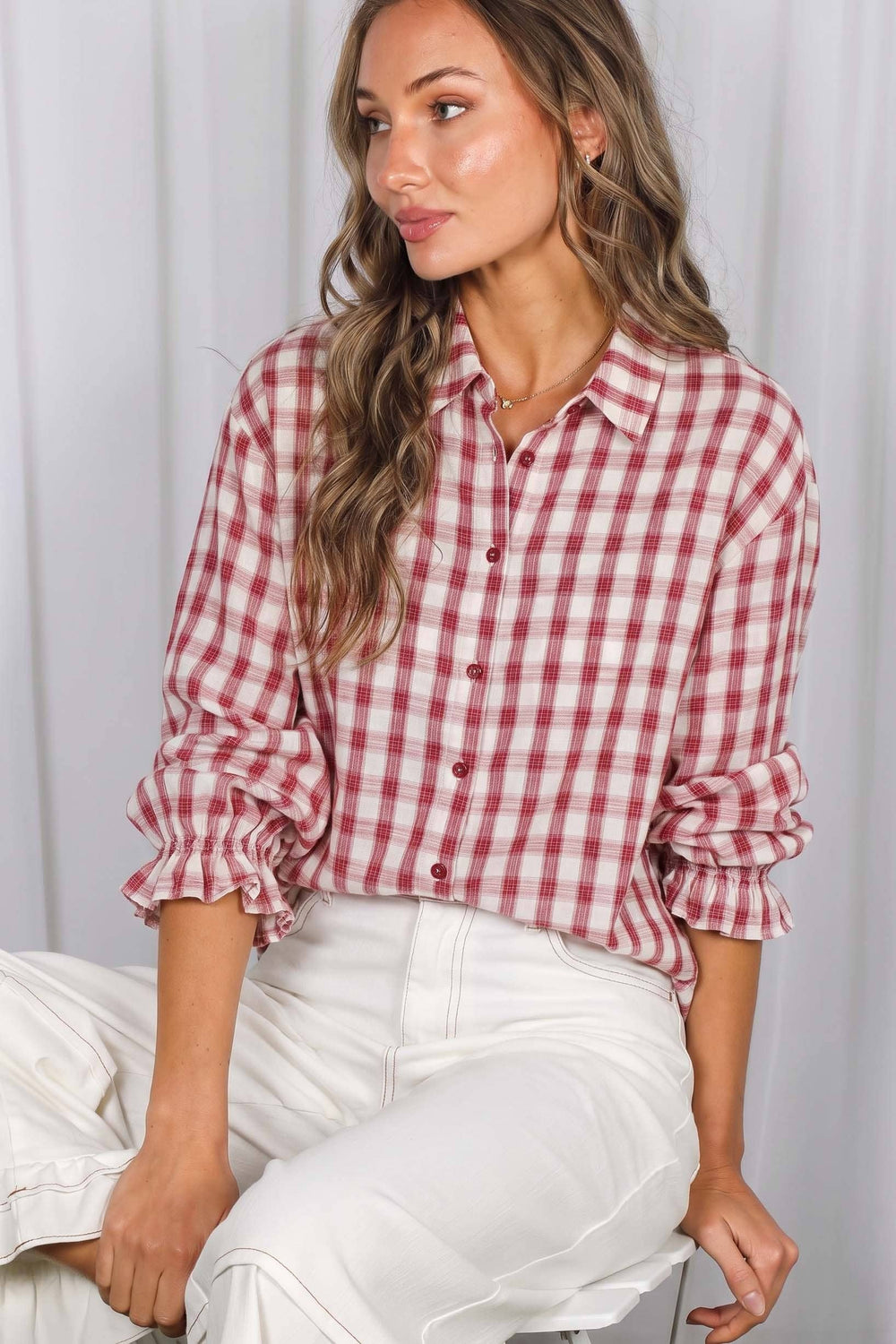 A woman wearing a red and white gingham button-up shirt with rolled-up sleeves and white pants, seated against a light gray curtain background.