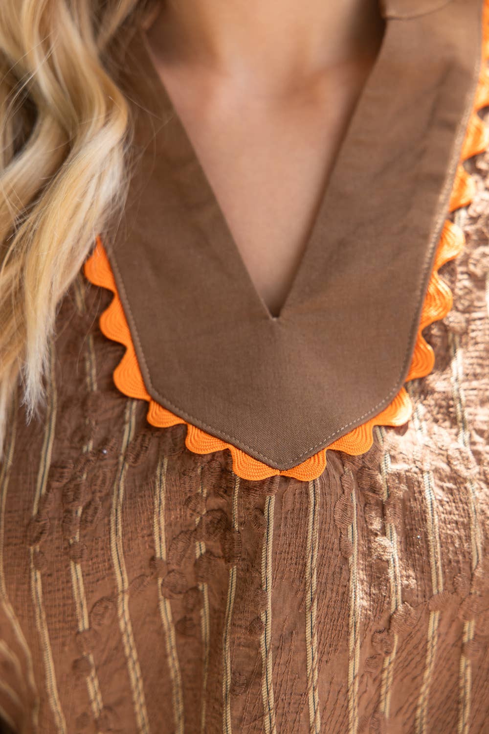 Close-up of a brown textured top with a wide brown V-neck collar trimmed in bright orange scalloped edges, worn by a model.