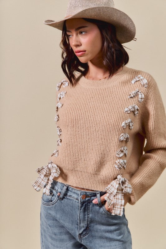 Woman wearing a beige sweater with bow details, blue jeans, and a straw hat on a beige background