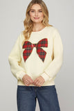 Woman wearing a cream sweater with a red tartan bow design on a plain background