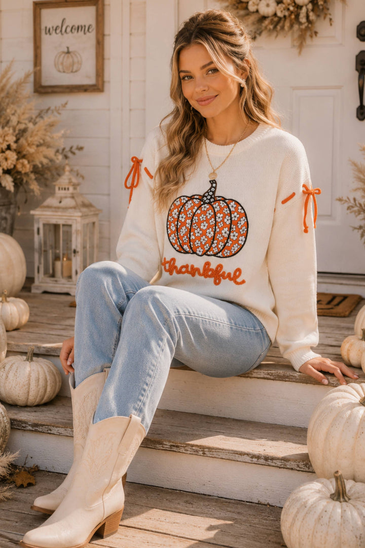 Woman wearing a white sweater with a pumpkin design sitting on steps surrounded by pumpkins.