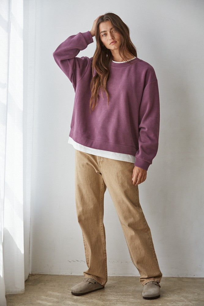 Woman wearing a purple sweater and beige pants standing against a white wall.