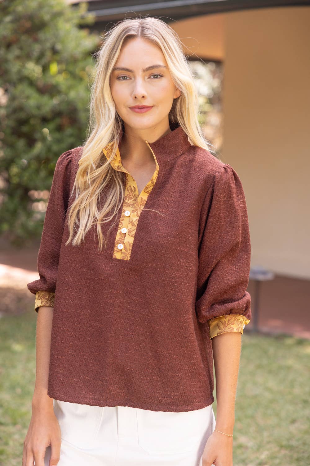 A woman wearing a rust-brown linen long-sleeve top with a gold patterned mandarin collar and cuffs, paired with white pants, standing outdoors in front of a house.