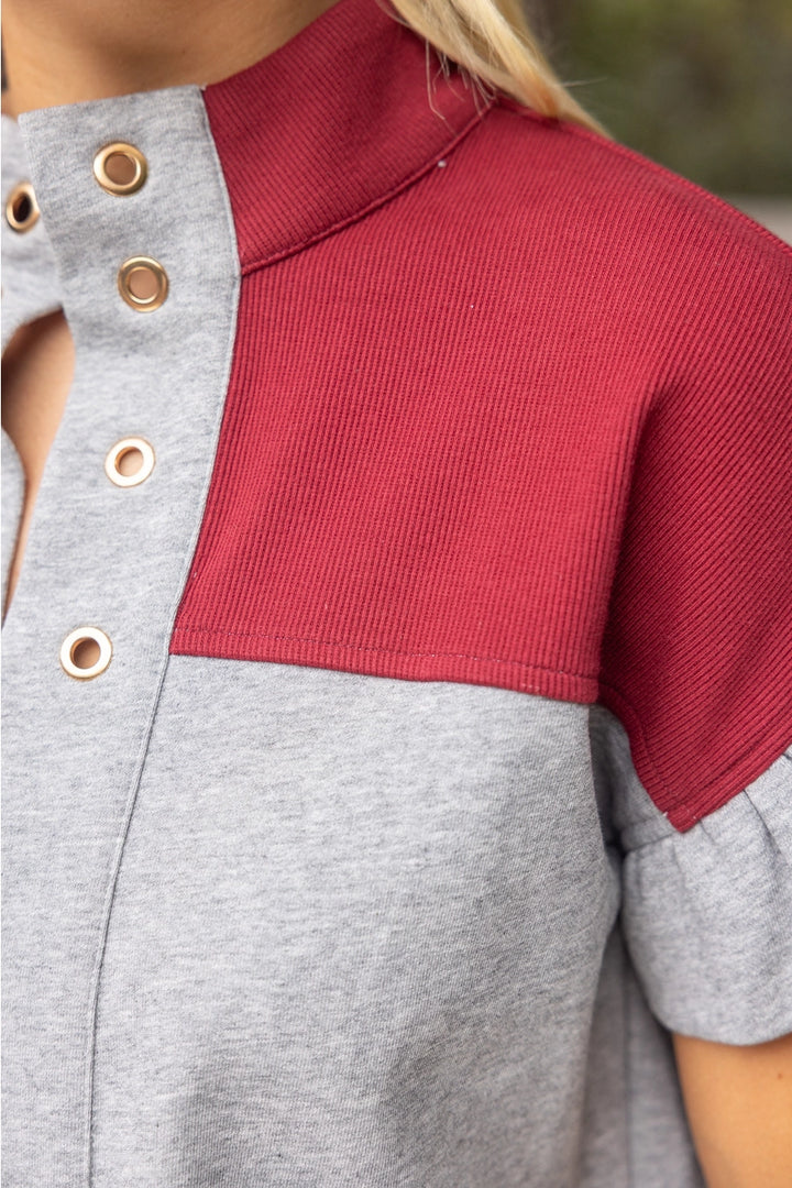 Close-up of a color-block short-sleeve top with a heather gray body and a contrasting dark red ribbed yoke, featuring gold-tone metal grommets on the front placket.