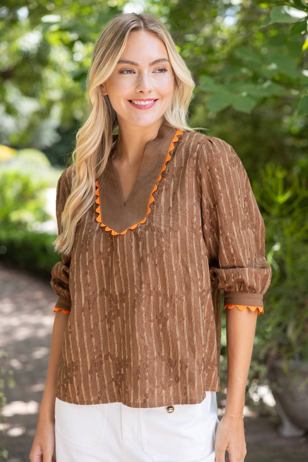A woman wearing a brown textured blouse with orange scalloped trim on the neckline and cuffs, paired with white pants, standing outdoors in front of a blurred green garden background.