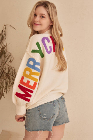 Person wearing a cream sweater with colorful letters spelling 'MERRY CHRISTMAS' on a beige background.