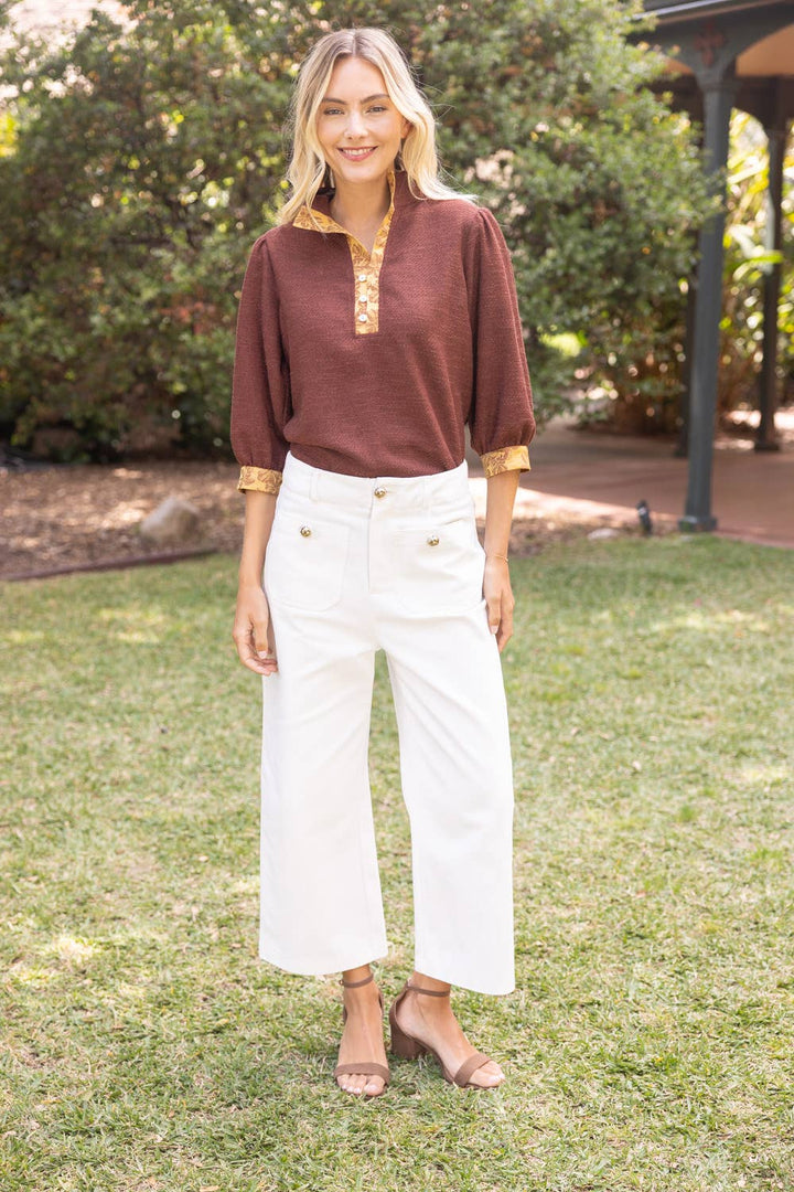 A woman standing outdoors on a grassy lawn wearing a rust-brown long-sleeve top with gold trim and white cropped pants.