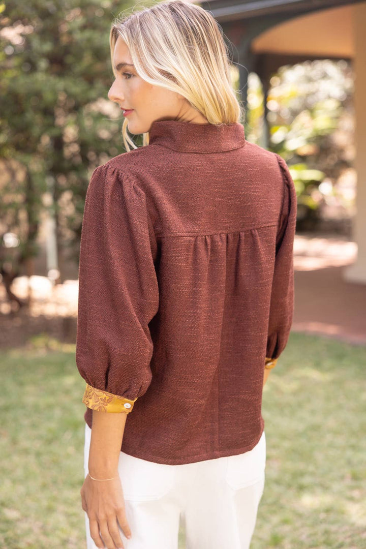 A woman shown from the back wearing a rust-brown long-sleeve blouse with gathered puff sleeves and white pants, standing outdoors on grass.