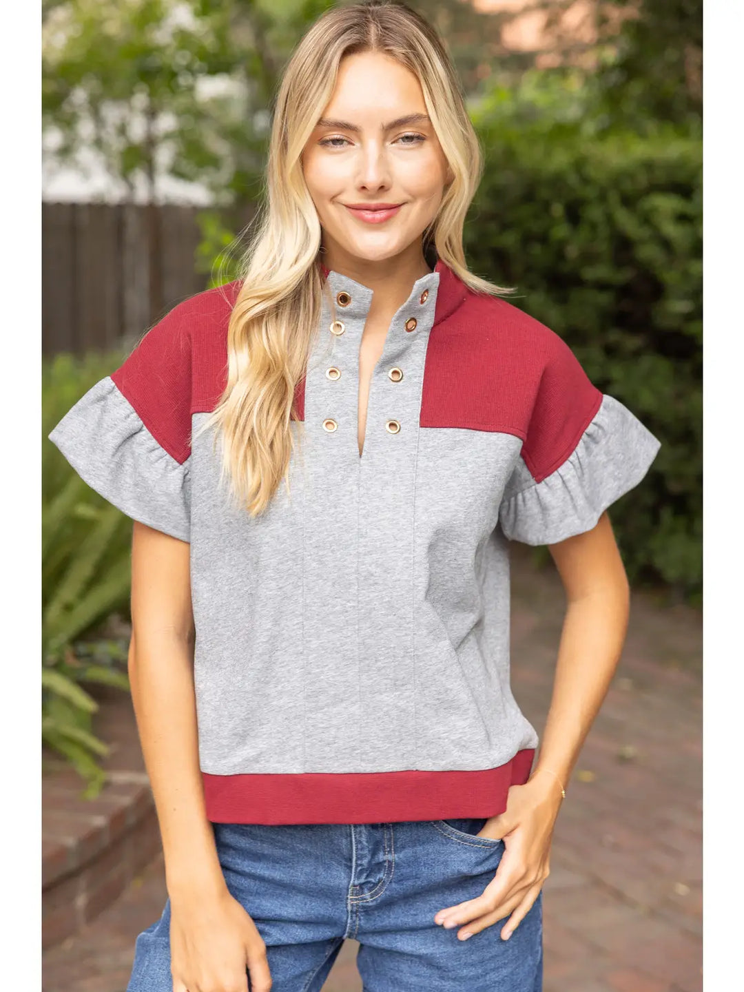 A woman wearing a color-block short-sleeve top with a heather gray body and sleeves, and a deep red yoke and hem, paired with blue jeans, set against a blurred outdoor background.