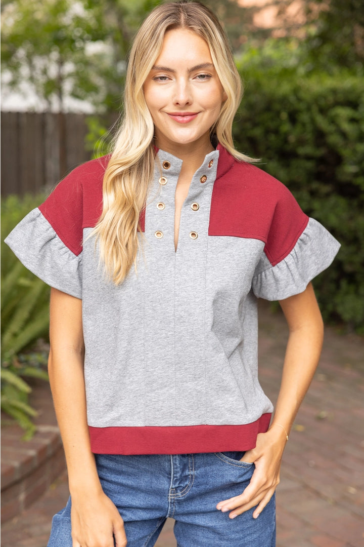 A woman wearing a color-block short-sleeve top with a heather gray body, burgundy yoke and hem, and ruffled sleeves, paired with blue jeans, standing outdoors.