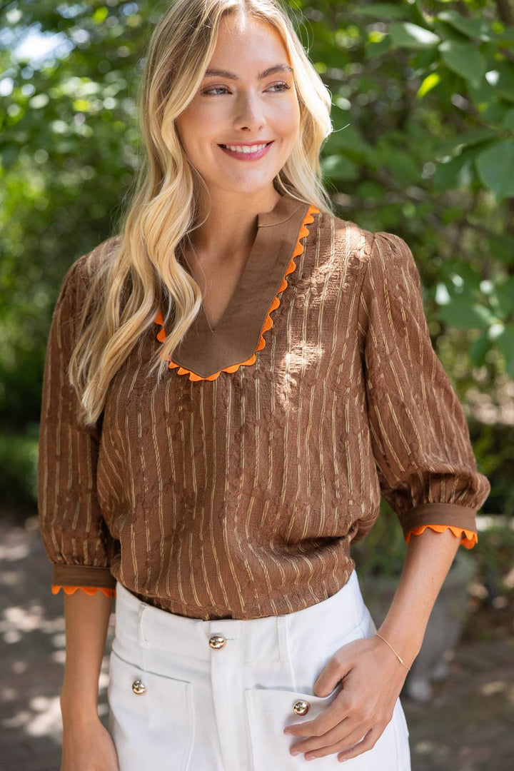 A woman wearing a brown pinstriped blouse with orange scalloped trim on the neckline and cuffs, paired with white pants, standing outdoors in front of a green leafy background.
