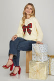 Woman wearing a cream sweater with a red tartan bow design, sitting on gift boxes against a plain background.