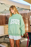 Person wearing a light green sweatshirt with 'JESUS' printed on it, standing outdoors.