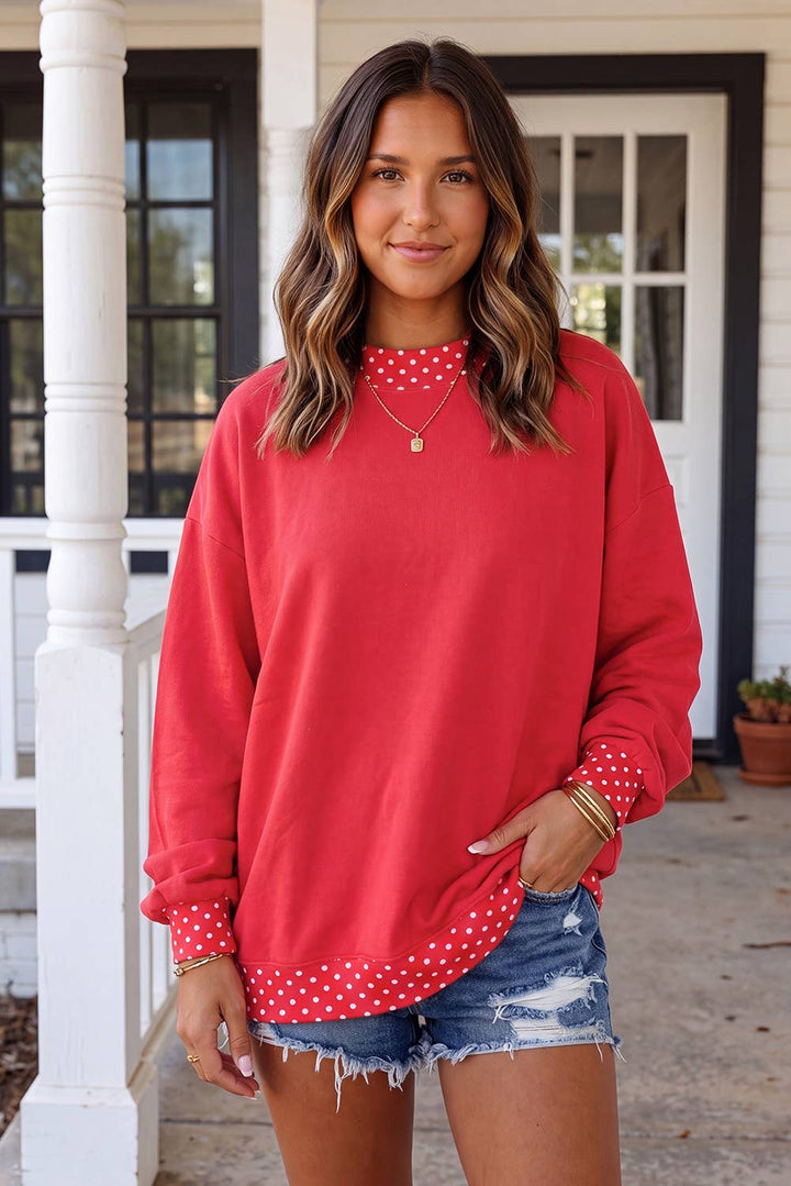 A woman wearing a coral red long-sleeve sweatshirt with white polka dot trim at the crew neck and hem, paired with denim shorts, standing on a porch.