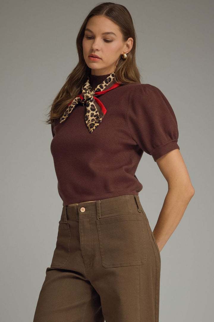 Woman wearing a brown top and high-waisted pants with a leopard print scarf on a gray background