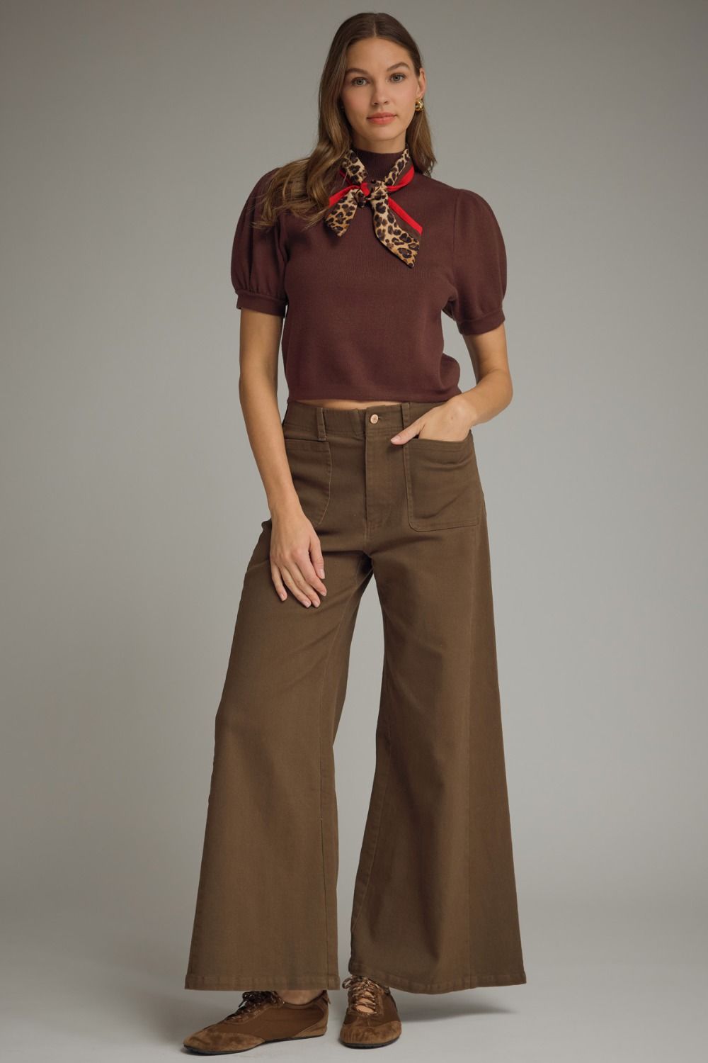 Woman wearing a brown top and wide-leg pants with a scarf, standing against a plain background