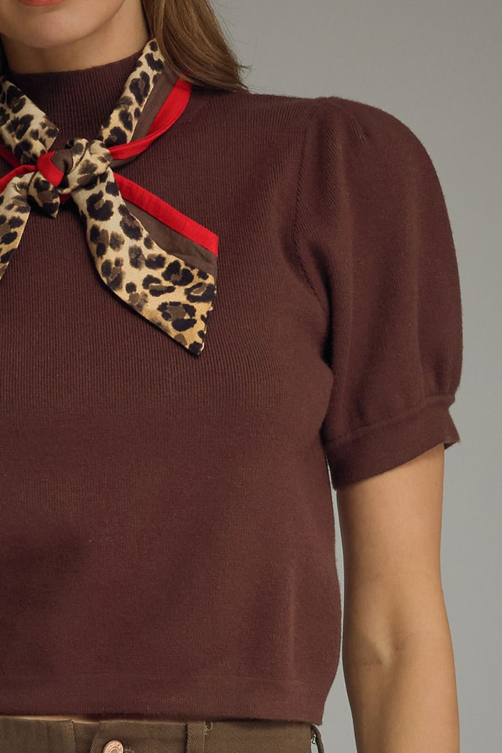 A close-up of a model wearing a brown ribbed knit short-sleeve top with puff sleeves and a leopard-print neck scarf tied in a bow, featuring red ribbon accents.