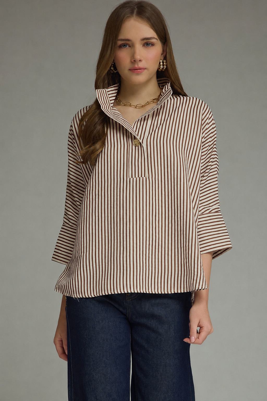 A woman modeling a brown and beige vertically striped button-up shirt with three-quarter sleeves, paired with dark blue jeans, against a solid gray studio background.