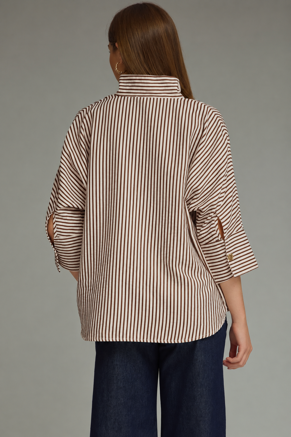 A woman viewed from the back wearing a beige and brown vertically striped button-down shirt with three-quarter sleeves and dark blue wide-leg jeans, set against a solid gray studio background.