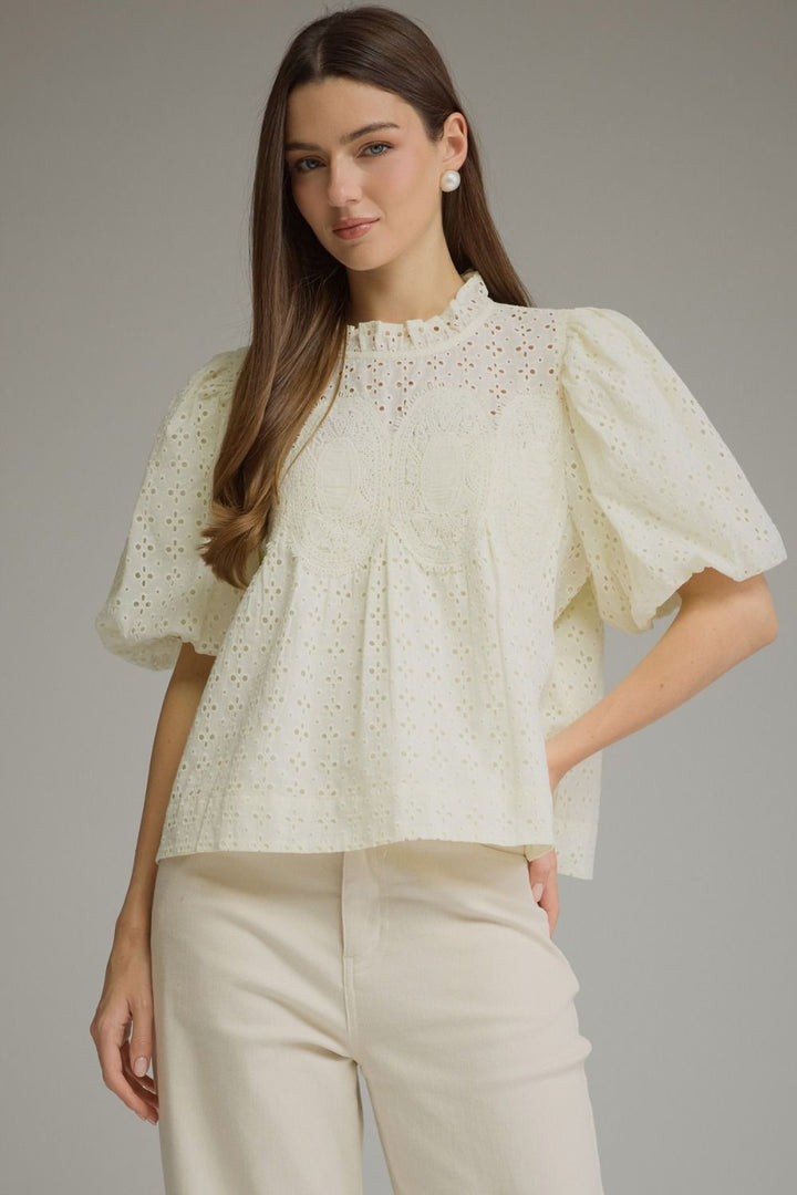 A woman modeling a cream-colored eyelet blouse with puff sleeves and a ruffled high neck, paired with cream trousers, against a solid gray studio background.