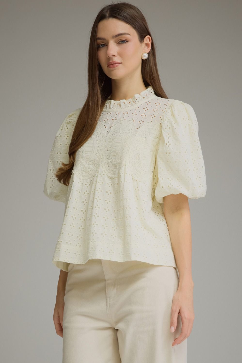 A woman modeling a cream-colored eyelet blouse with puff sleeves and a ruffled high neckline, paired with matching cream trousers, against a solid gray studio background.