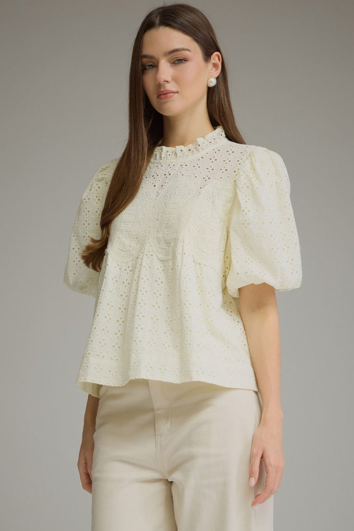 A woman modeling a cream-colored eyelet blouse with puff sleeves and a ruffled high neckline, paired with matching cream trousers, against a solid gray studio background.